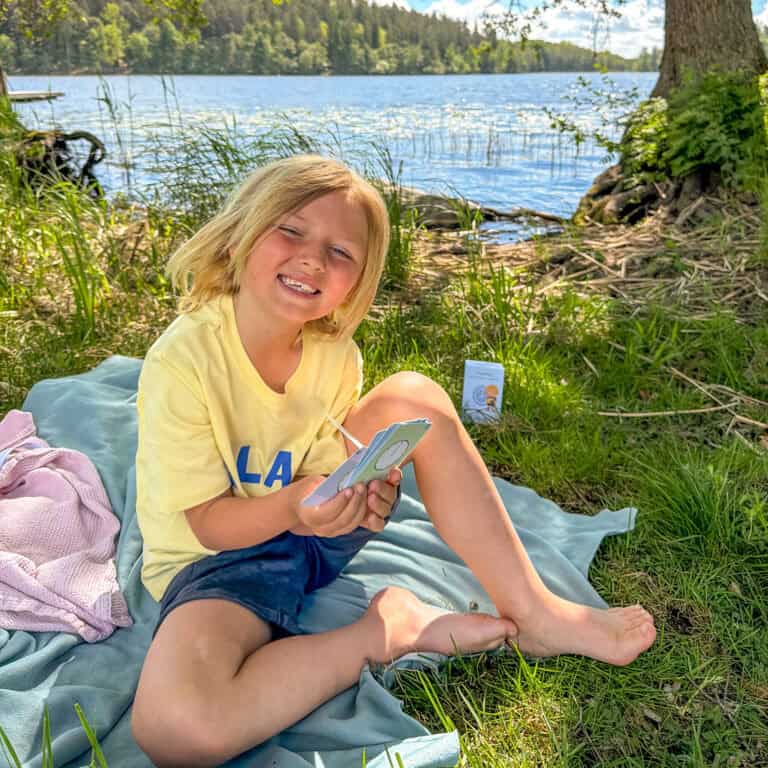 En glad pojke i sommarkläder sitter på en filt vid sjön och klurar med Klures kortlekar och lär sig klockan.